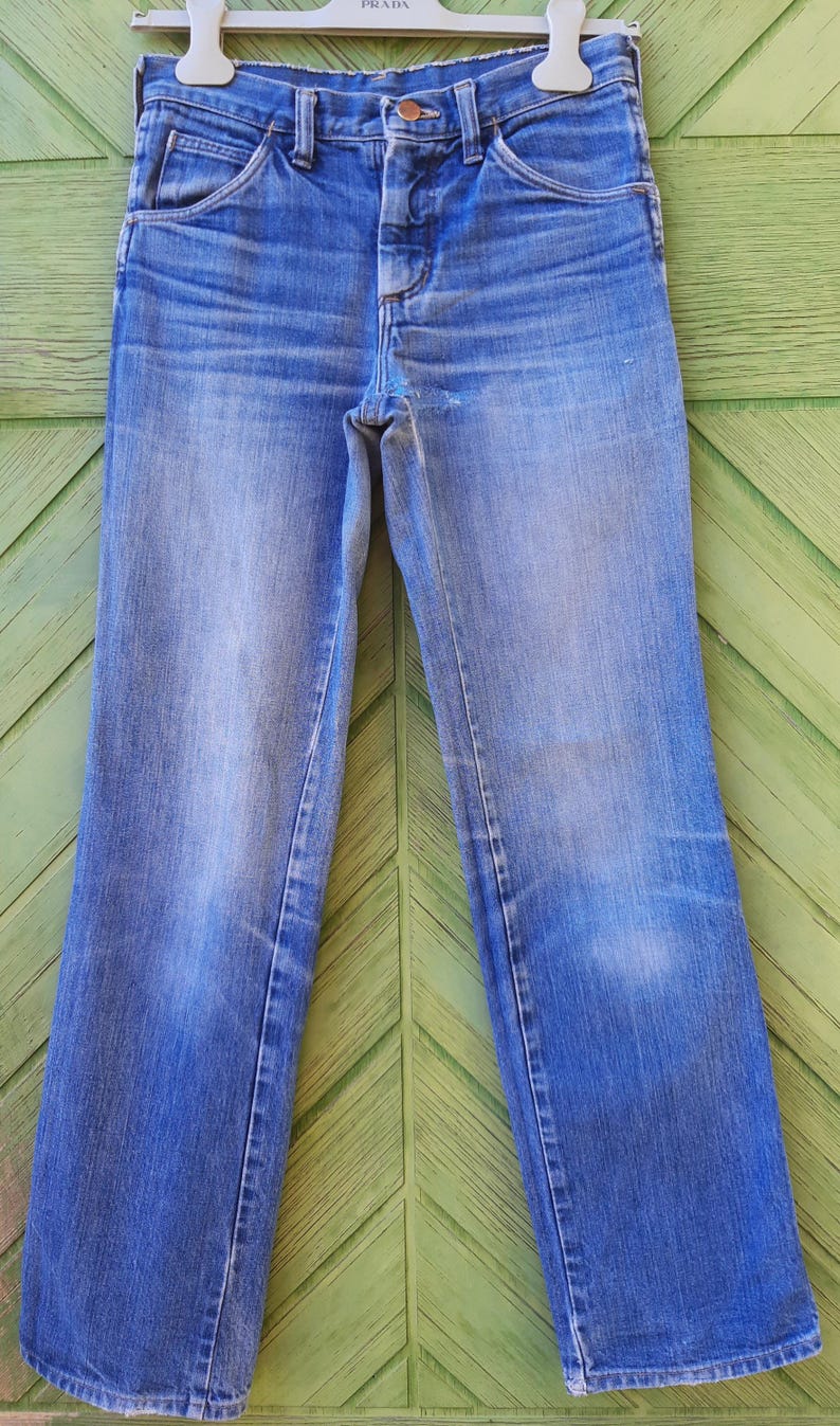 May include: A pair of light blue denim jeans with a copper button and a small pocket on the right side. The jeans are hanging on a white hanger with a "PRADA" label.