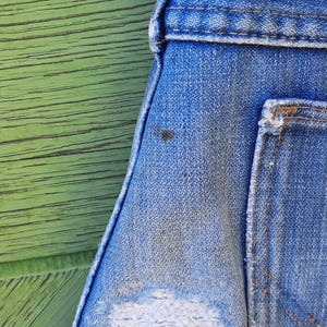 Pode incluir: Close-up de macac&atilde;o jeans azul com um visual desgastado e usado. O jeans tem uma apar&ecirc;ncia desbotada com um grande remendo branco desfiado. O fundo &eacute; uma superf&iacute;cie de madeira verde desgastada.