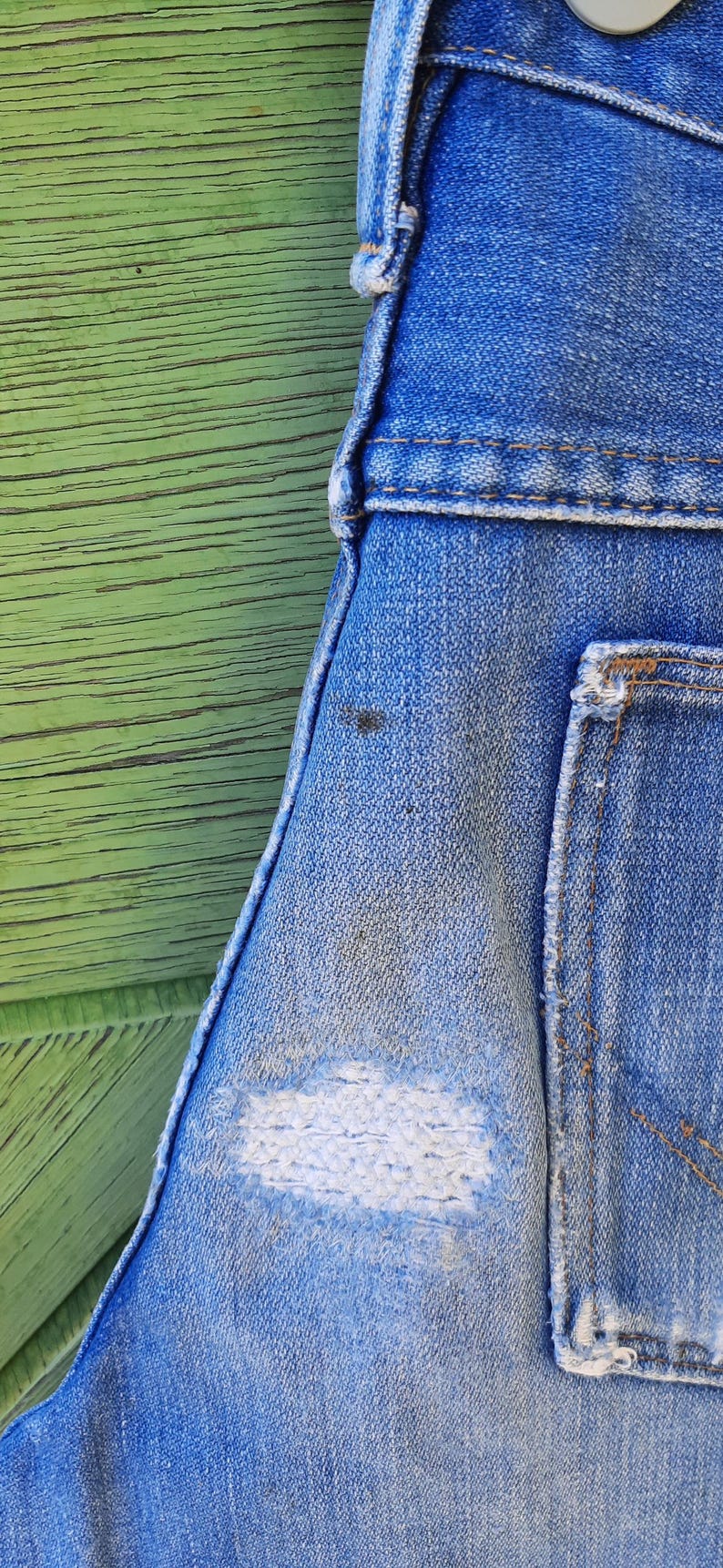 Pode incluir: Close-up de macac&atilde;o jeans azul com um visual desgastado e usado. O jeans tem uma apar&ecirc;ncia desbotada com um grande remendo branco desfiado. O fundo &eacute; uma superf&iacute;cie de madeira verde desgastada.