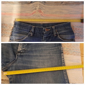 May include: Close-up of blue denim jeans with a button closure and pockets. A yellow measuring tape is laid across the waistband, and another measures the leg length. The jeans are on a patterned rug.