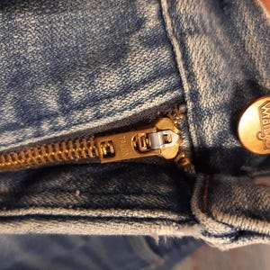 May include: Close-up of a light blue denim garment, featuring a brass-colored Talon zipper and a Wrangler button. The zipper is partially open, revealing the teeth and slider. The Wrangler button is round with the brand name embossed.