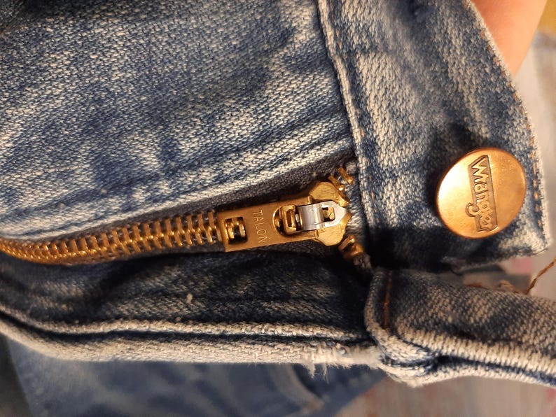 May include: Close-up of a light blue denim garment, featuring a brass-colored Talon zipper and a Wrangler button. The zipper is partially open, revealing the teeth and slider. The Wrangler button is round with the brand name embossed.