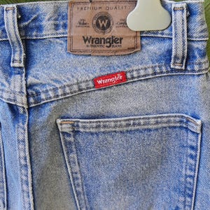 May include: Light blue Wrangler jeans with a classic design. The back of the jeans features a pocket and a brown leather patch with the Wrangler logo. A red Wrangler tag is sewn on the back pocket.