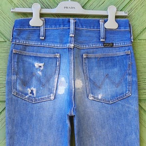 Pode incluir: Jeans de ganga azul desbotada com detalhes desgastados, incluindo rasgos e marcas de uso. Os bolsos traseiros apresentam as costuras do log&oacute;tipo Wrangler. Os jeans est&atilde;o pendurados num cabide Prada branco.