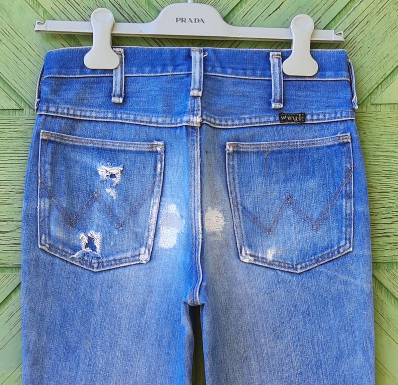 Pode incluir: Jeans de ganga azul desbotada com detalhes desgastados, incluindo rasgos e marcas de uso. Os bolsos traseiros apresentam as costuras do log&oacute;tipo Wrangler. Os jeans est&atilde;o pendurados num cabide Prada branco.
