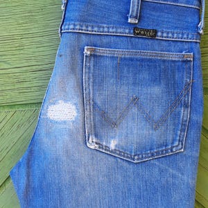 Pode incluir: Cal&ccedil;as de ganga azuis com lavagem desbotada e um remendo desgastado na coxa esquerda. As cal&ccedil;as de ganga apresentam um design cl&aacute;ssico de cinco bolsos, um bolso traseiro com um design 'W' costurado e uma etiqueta 'Wrangler'.