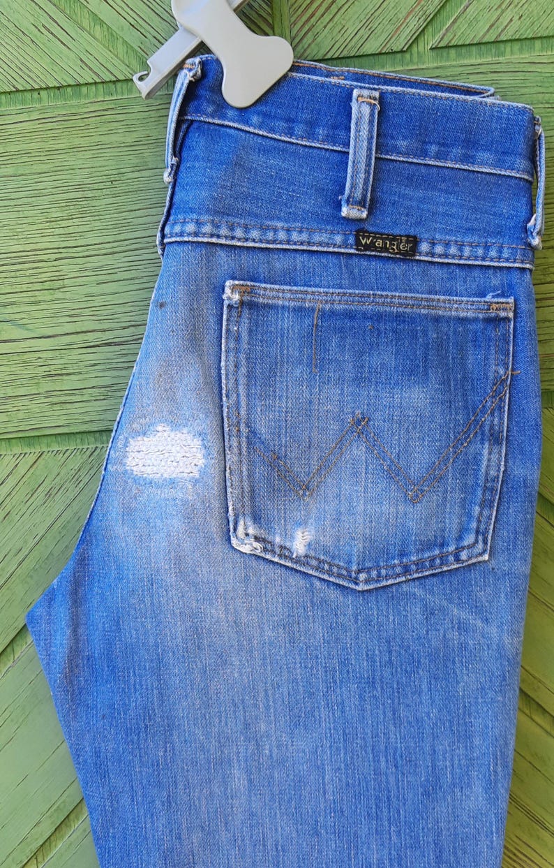 Pode incluir: Cal&ccedil;as de ganga azuis com lavagem desbotada e um remendo desgastado na coxa esquerda. As cal&ccedil;as de ganga apresentam um design cl&aacute;ssico de cinco bolsos, um bolso traseiro com um design 'W' costurado e uma etiqueta 'Wrangler'.