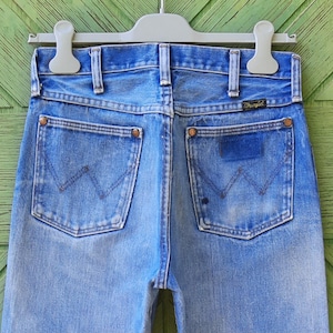 May include: Light blue denim jeans featuring a high-waisted design with belt loops, a Wrangler brand tag on the back pocket, and copper-toned rivets. The back pockets display a signature W-shaped stitching pattern in a faded, worn-in wash.