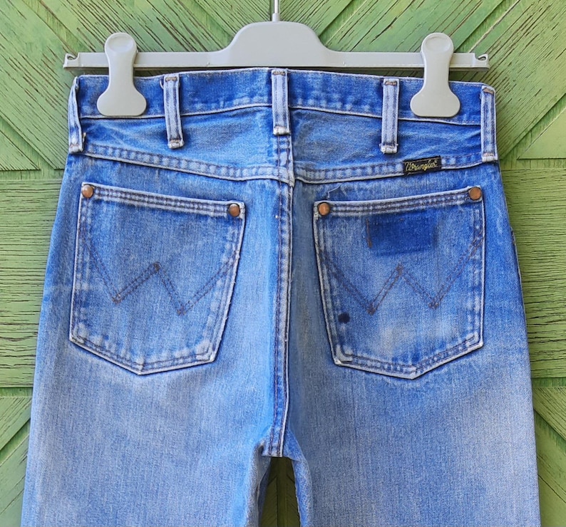 May include: Light blue denim jeans featuring a high-waisted design with belt loops, a Wrangler brand tag on the back pocket, and copper-toned rivets. The back pockets display a signature W-shaped stitching pattern in a faded, worn-in wash.
