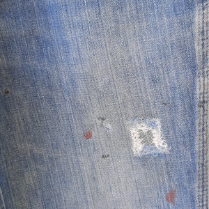 Pode incluir: Close-up de jeans de ganga azul clara com um aspeto desgastado e usado. Apresenta um remendo vis&iacute;vel e v&aacute;rias pequenas marcas vermelhas e pretas, adicionando &agrave; est&eacute;tica vintage. O tecido mostra gr&atilde;o vertical.