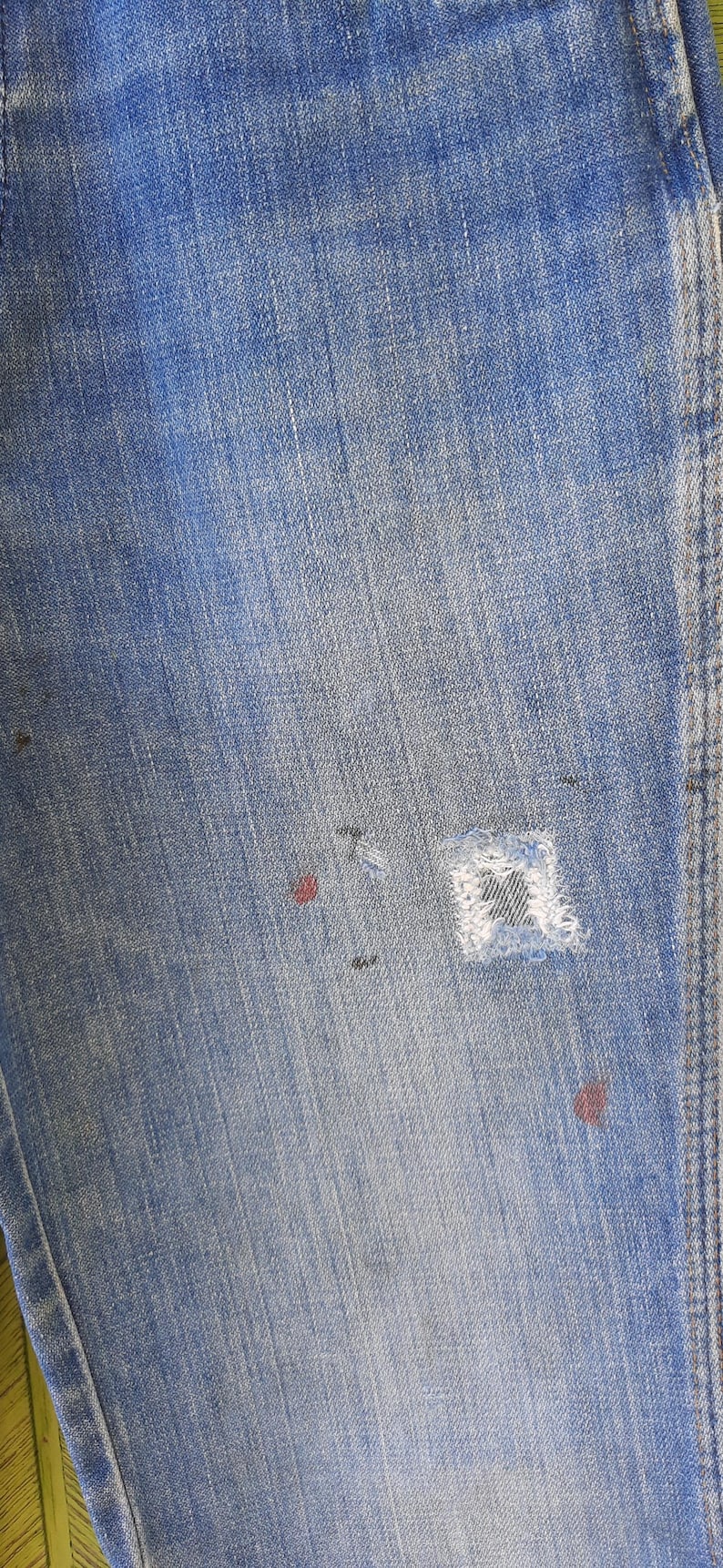 Pode incluir: Close-up de jeans de ganga azul clara com um aspeto desgastado e usado. Apresenta um remendo vis&iacute;vel e v&aacute;rias pequenas marcas vermelhas e pretas, adicionando &agrave; est&eacute;tica vintage. O tecido mostra gr&atilde;o vertical.