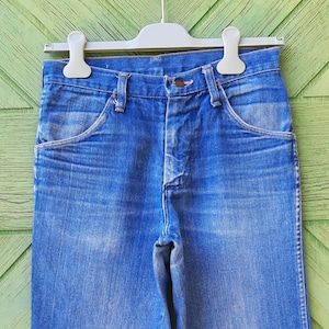 May include: Close-up of blue denim jeans hanging on a white hanger. The jeans have a classic five-pocket design with a button and zipper closure. The denim has a faded, worn look.