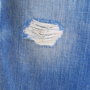 Pode incluir: Close-up de tecido denim azul com um detalhe rasgado e desgastado. A rasgadura revela fios brancos, criando um contraste texturizado com o denim azul. A imagem destaca a est&eacute;tica desgastada e casual dos jeans.