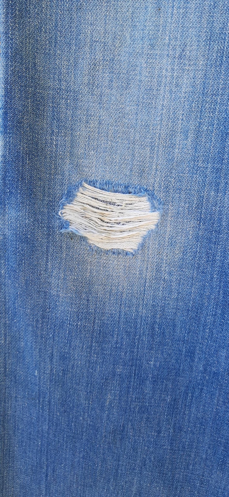 Pode incluir: Close-up de tecido denim azul com um detalhe rasgado e desgastado. A rasgadura revela fios brancos, criando um contraste texturizado com o denim azul. A imagem destaca a est&eacute;tica desgastada e casual dos jeans.
