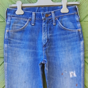 Pode incluir: Jeans de ganga azul desbotada pendurados num cabide branco. Os jeans t&ecirc;m um design cl&aacute;ssico de cinco bolsos, fecho de bot&atilde;o e z&iacute;per e presilhas para cinto. H&aacute; salpicos de tinta na frente dos jeans.