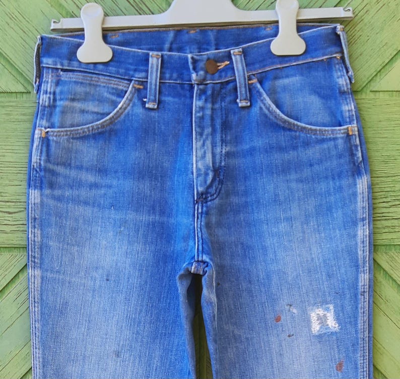 Pode incluir: Jeans de ganga azul desbotada pendurados num cabide branco. Os jeans t&ecirc;m um design cl&aacute;ssico de cinco bolsos, fecho de bot&atilde;o e z&iacute;per e presilhas para cinto. H&aacute; salpicos de tinta na frente dos jeans.
