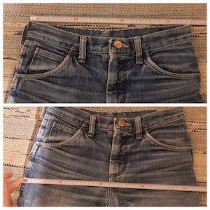May include: Two pairs of blue denim jeans with a copper button and a single pocket on each side. The jeans are laid flat on a woven surface and measured with a tape measure.