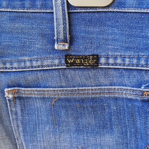 Pode incluir: Close-up de cal&ccedil;as de ganga azuis com um bolso traseiro e a etiqueta da marca Wrangler. As cal&ccedil;as de ganga est&atilde;o penduradas num cabide branco. O denim tem um aspeto desgastado.