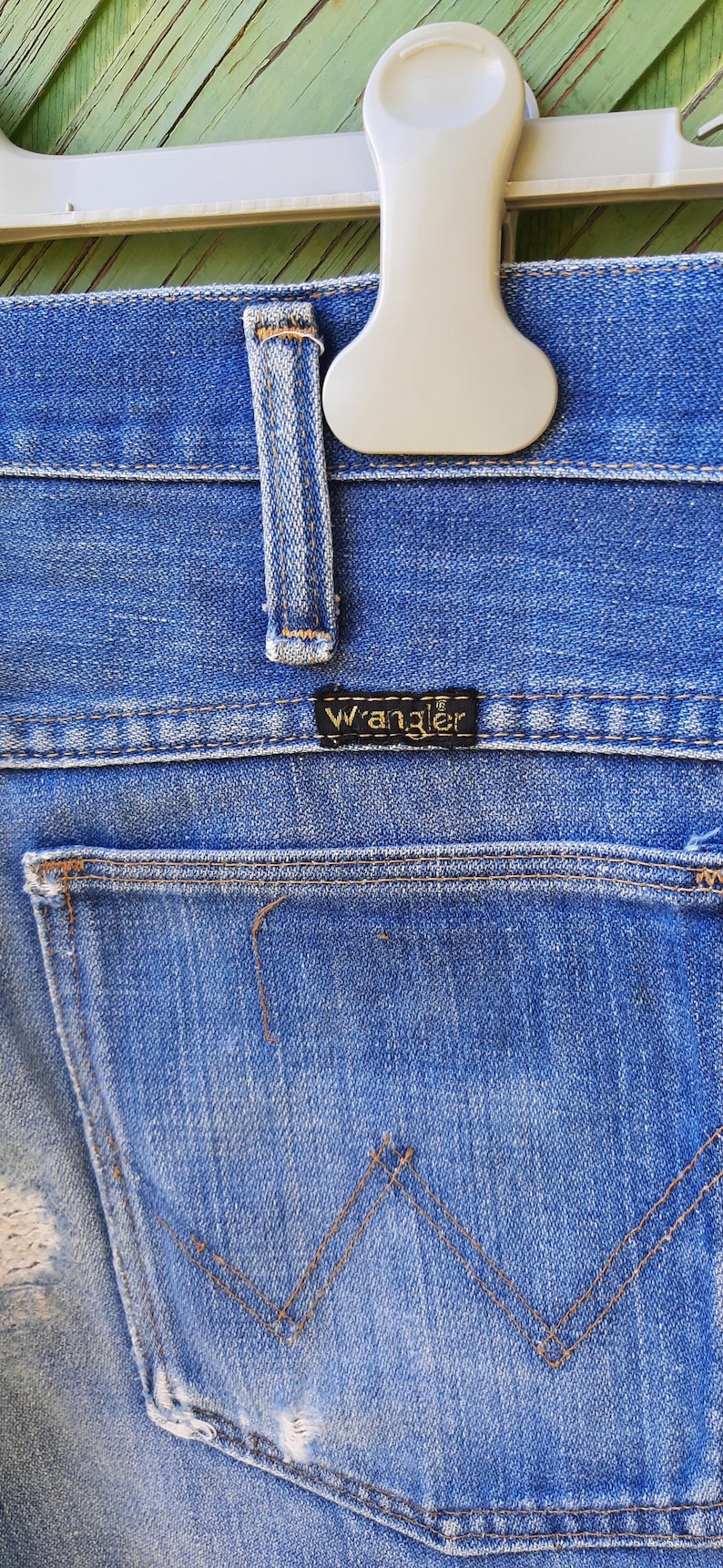 Pode incluir: Close-up de cal&ccedil;as de ganga azuis com um bolso traseiro e a etiqueta da marca Wrangler. As cal&ccedil;as de ganga est&atilde;o penduradas num cabide branco. O denim tem um aspeto desgastado.