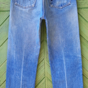 May include: A pair of light blue denim jeans featuring a high-waisted cut, two back pockets with a signature W-shaped stitch pattern, copper-toned rivets, and a small Wrangler brand label on the right pocket, all suspended from a plastic hanger.
