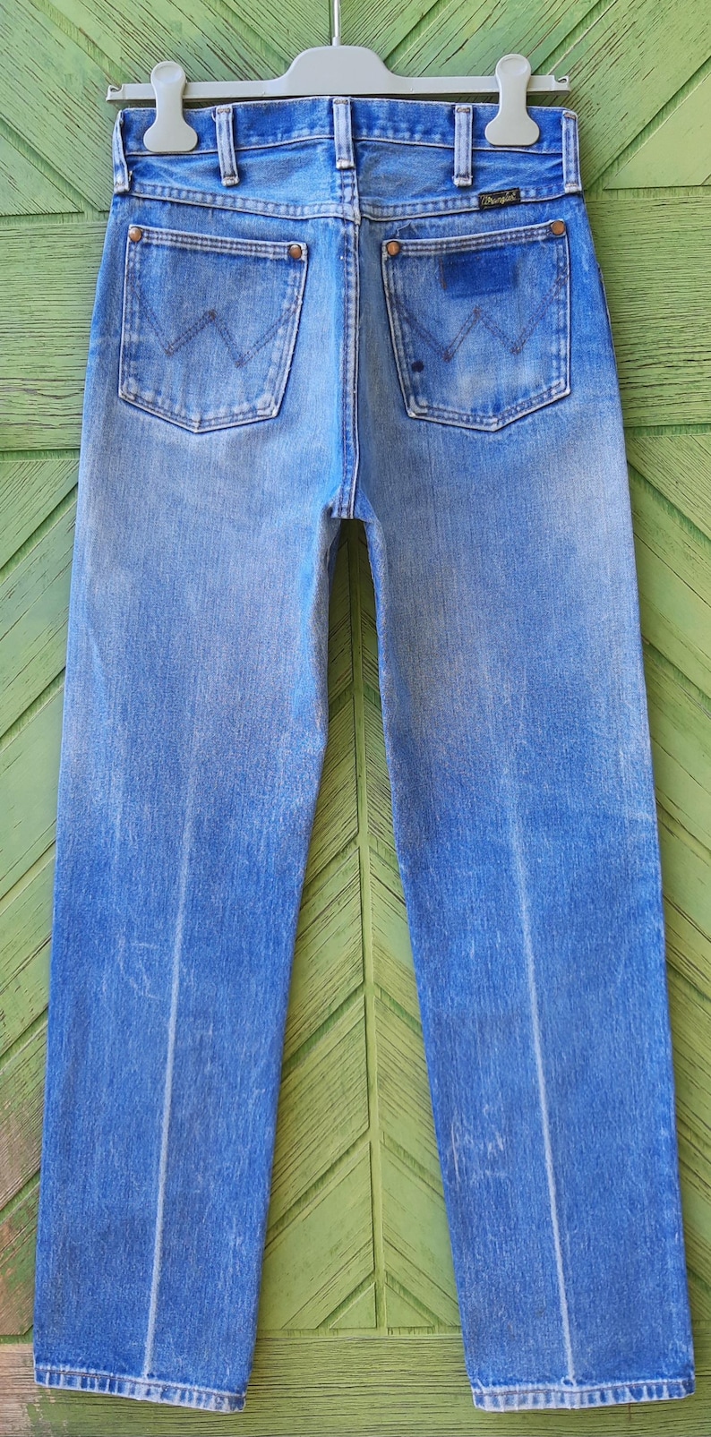 May include: A pair of light blue denim jeans featuring a high-waisted cut, two back pockets with a signature W-shaped stitch pattern, copper-toned rivets, and a small Wrangler brand label on the right pocket, all suspended from a plastic hanger.