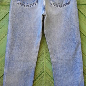 May include: Light blue denim jeans with a classic straight leg fit. The jeans feature two back pockets and a Wrangler brand patch. The denim has a faded wash, creating a vintage look. The jeans are hung on a hanger.