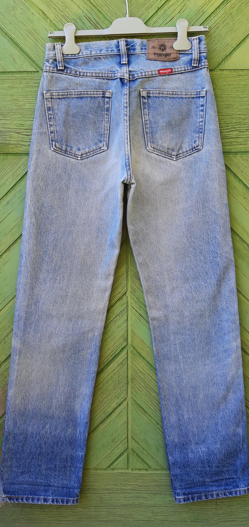 May include: Light blue denim jeans with a classic straight leg fit. The jeans feature two back pockets and a Wrangler brand patch. The denim has a faded wash, creating a vintage look. The jeans are hung on a hanger.