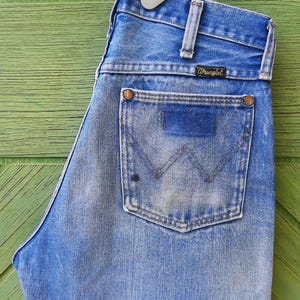 May include: A pair of light blue denim jeans featuring a high-waisted design with a classic five-pocket construction. The back pocket displays a stitched W pattern and a black Wrangler brand label. The fabric shows a faded wash with copper rivets.