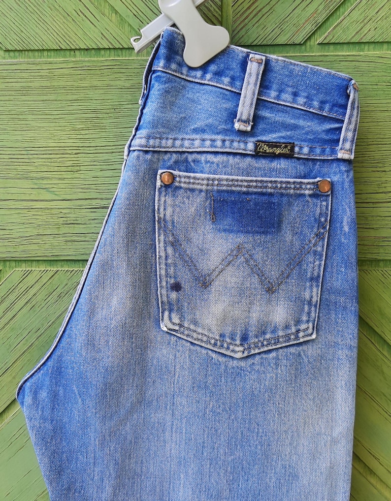 May include: A pair of light blue denim jeans featuring a high-waisted design with a classic five-pocket construction. The back pocket displays a stitched W pattern and a black Wrangler brand label. The fabric shows a faded wash with copper rivets.