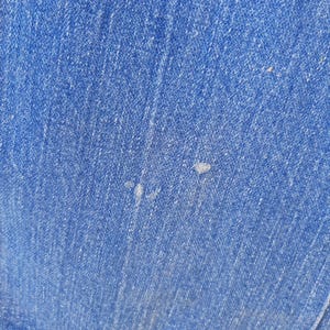 May include: Close-up of blue denim fabric. The denim has a textured appearance with visible weave and slight variations in color. There are a few small, light-colored spots on the fabric. The bottom edge of the fabric is visible, showing a seam.