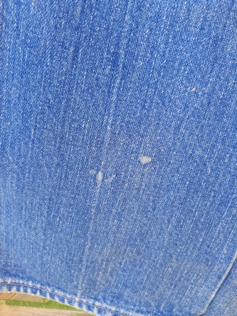 May include: Close-up of blue denim fabric. The denim has a textured appearance with visible weave and slight variations in color. There are a few small, light-colored spots on the fabric. The bottom edge of the fabric is visible, showing a seam.