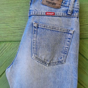 May include: Light blue Wrangler jeans with a classic five-pocket design. The jeans feature a brown leather patch with the Wrangler logo and a red Wrangler tag on the back pocket. The jeans are made of denim.