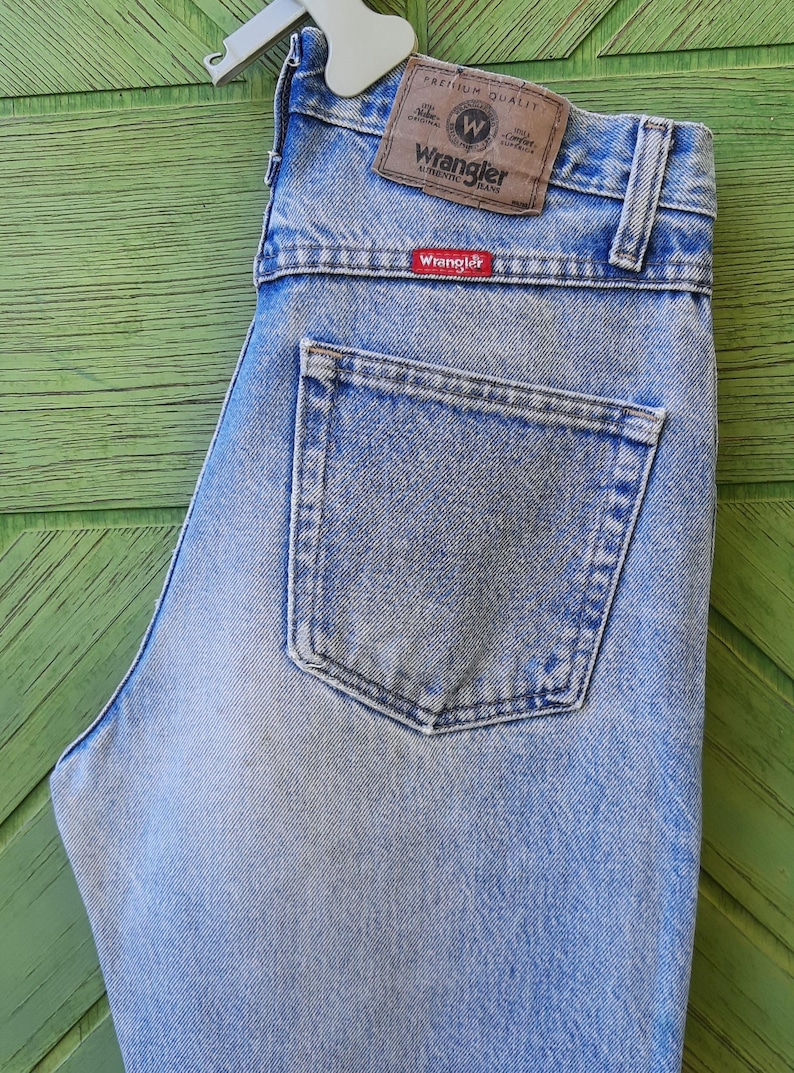 May include: Light blue Wrangler jeans with a classic five-pocket design. The jeans feature a brown leather patch with the Wrangler logo and a red Wrangler tag on the back pocket. The jeans are made of denim.
