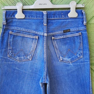 May include: Close-up of the back of blue denim jeans with two pockets and a Wrangler label. The jeans are hanging on a white hanger with the word "PRADA" printed on it. The denim has a faded appearance.