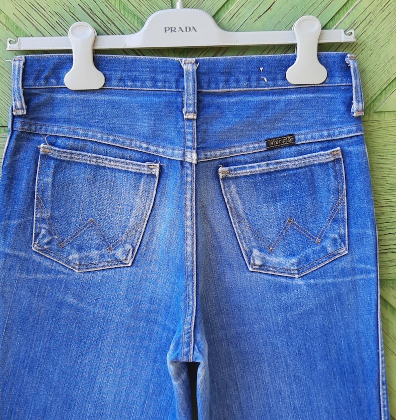 May include: Close-up of the back of blue denim jeans with two pockets and a Wrangler label. The jeans are hanging on a white hanger with the word "PRADA" printed on it. The denim has a faded appearance.