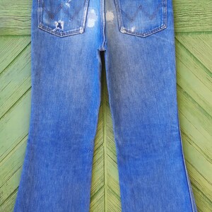 Pode incluir: Jeans vintage azuis de ganga com um aspeto desgastado, com remendos gastos e bainhas desfiadas. Os jeans t&ecirc;m um design cl&aacute;ssico de cinco bolsos e uma perna flare. O log&oacute;tipo Wrangler &eacute; vis&iacute;vel no bolso de tr&aacute;s.