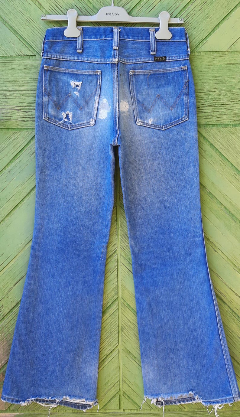 Pode incluir: Jeans vintage azuis de ganga com um aspeto desgastado, com remendos gastos e bainhas desfiadas. Os jeans t&ecirc;m um design cl&aacute;ssico de cinco bolsos e uma perna flare. O log&oacute;tipo Wrangler &eacute; vis&iacute;vel no bolso de tr&aacute;s.