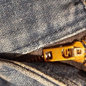 May include: Close-up of a denim garment with a gold-colored zipper. The zipper is partially open, revealing the teeth and the Talon brand name. A copper-colored button is visible.