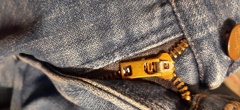 May include: Close-up of a denim garment with a gold-colored zipper. The zipper is partially open, revealing the teeth and the Talon brand name. A copper-colored button is visible.