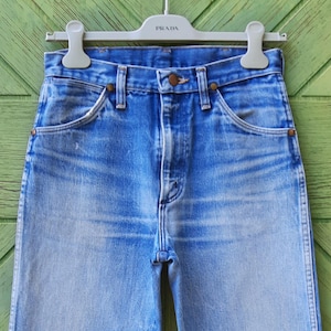 May include: A pair of high-waisted blue denim jeans featuring a faded wash, a button fly closure, and copper-toned rivets at the pocket corners. The jeans are suspended from a light gray plastic hanger with the brand name Prada embossed on it.