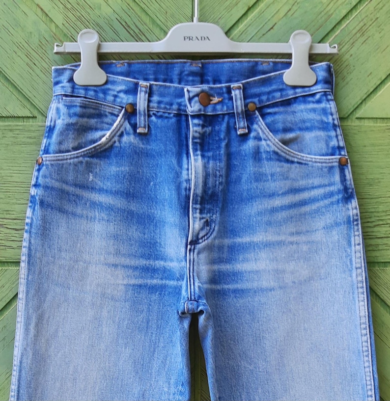 May include: A pair of high-waisted blue denim jeans featuring a faded wash, a button fly closure, and copper-toned rivets at the pocket corners. The jeans are suspended from a light gray plastic hanger with the brand name Prada embossed on it.