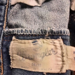 May include: A close-up of a blue denim jean with a white label that says "Wrangler" and "Made in Malta".