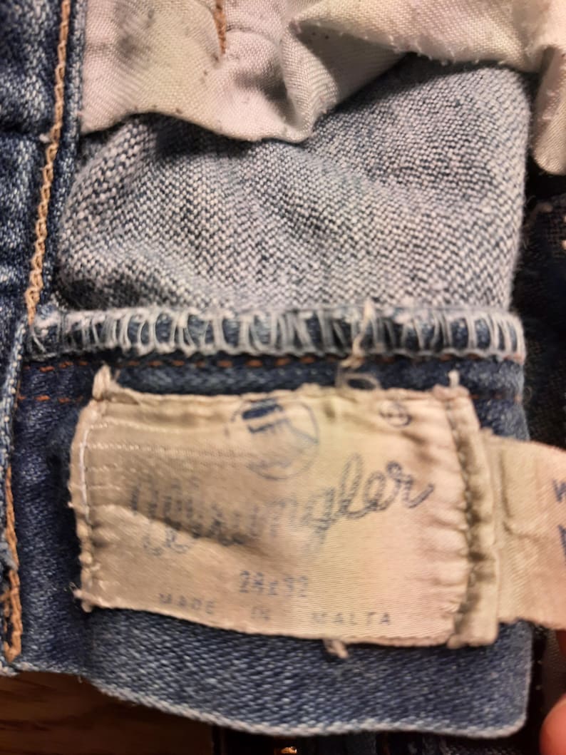 May include: A close-up of a blue denim jean with a white label that says "Wrangler" and "Made in Malta".