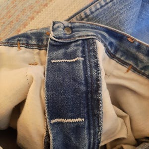May include: Close-up of the button and buttonhole area of blue denim jeans. The jeans have a metal button and white stitching. The interior lining is a light beige color. The fabric shows signs of wear.