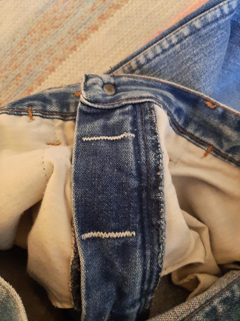 May include: Close-up of the button and buttonhole area of blue denim jeans. The jeans have a metal button and white stitching. The interior lining is a light beige color. The fabric shows signs of wear.
