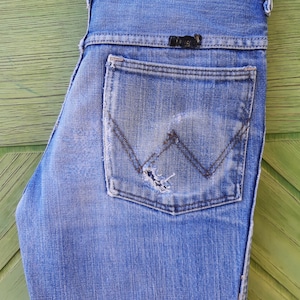 May include: A pair of blue denim jeans with a back pocket that has a distressed look and a small black leather label with the text "Wrangler" on it.