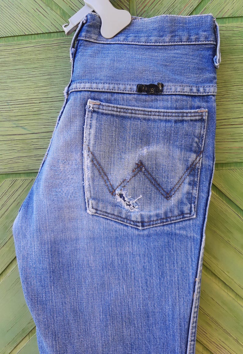 May include: A pair of blue denim jeans with a back pocket that has a distressed look and a small black leather label with the text "Wrangler" on it.