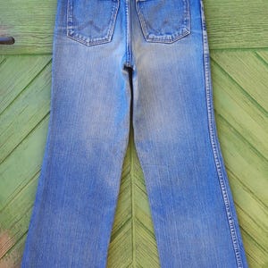 May include: Vintage blue denim Wrangler jeans with a high waist and flared legs. The jeans have two back pockets and a Wrangler logo. The jeans are hanging on a Prada hanger.