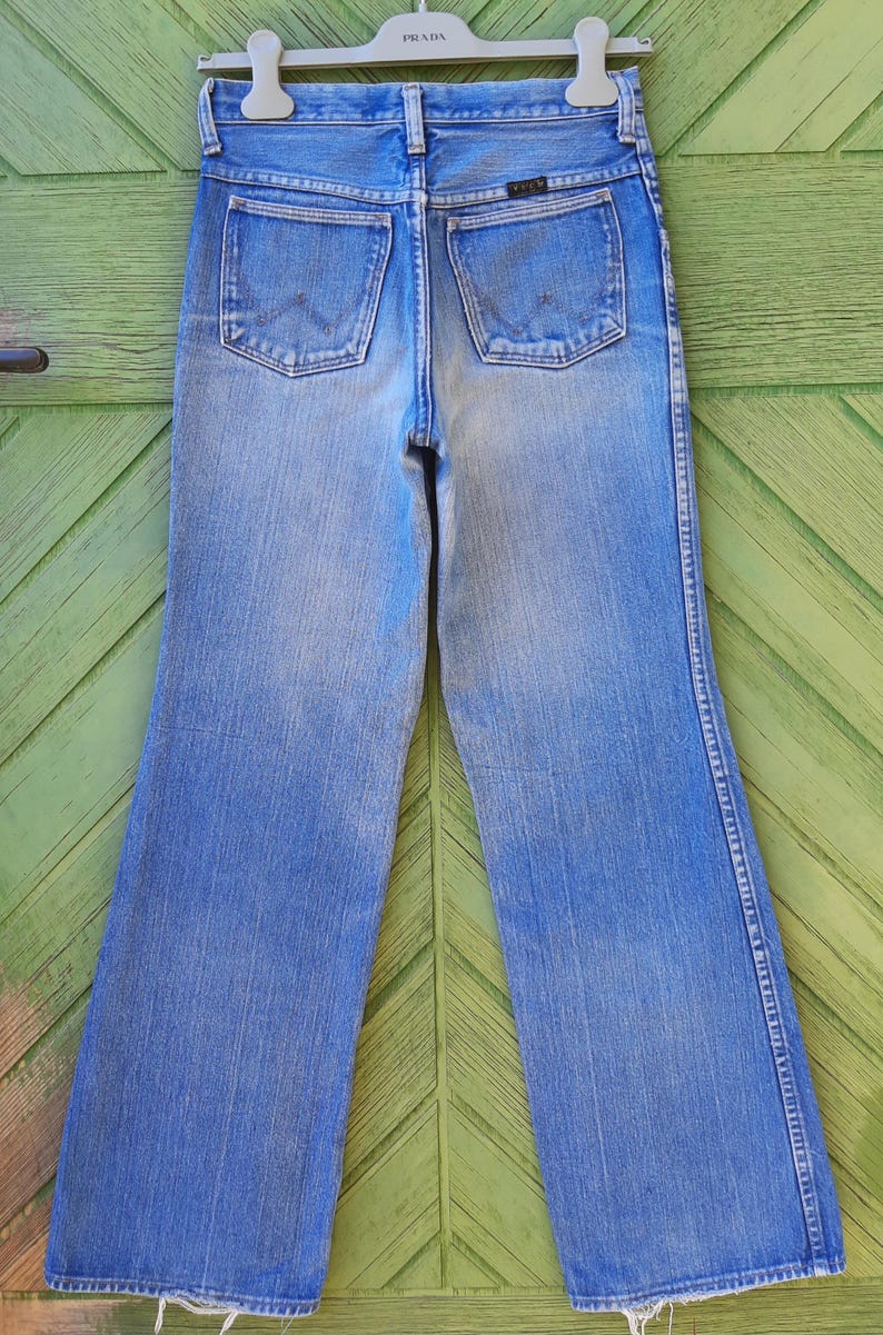 May include: Vintage blue denim Wrangler jeans with a high waist and flared legs. The jeans have two back pockets and a Wrangler logo. The jeans are hanging on a Prada hanger.