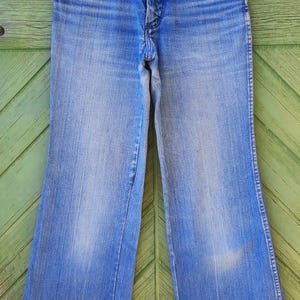 May include: Light blue denim jeans with a classic five-pocket design. The jeans have a high waist and a flared leg, with a faded wash and subtle wear marks. The jeans are hanging on a white hanger against a green background.